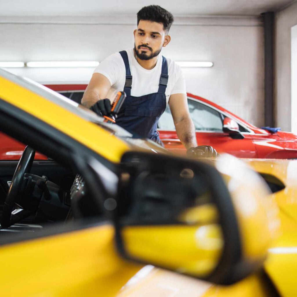 Anti rain coating at auto service. Male worker in blue overalls and protective gloves, on a professional car wash service, applying anti rain coating on a windshield of luxury yellow car.
