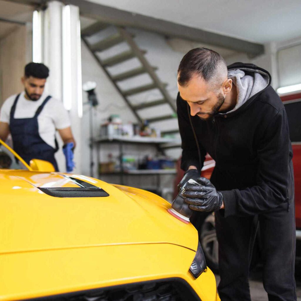 Auto detailing service, polishing of the car. Team of young men workers polishing yellow car body with orbital polisher and wax and wipe the windshield to eliminate contaminants from the surface.
